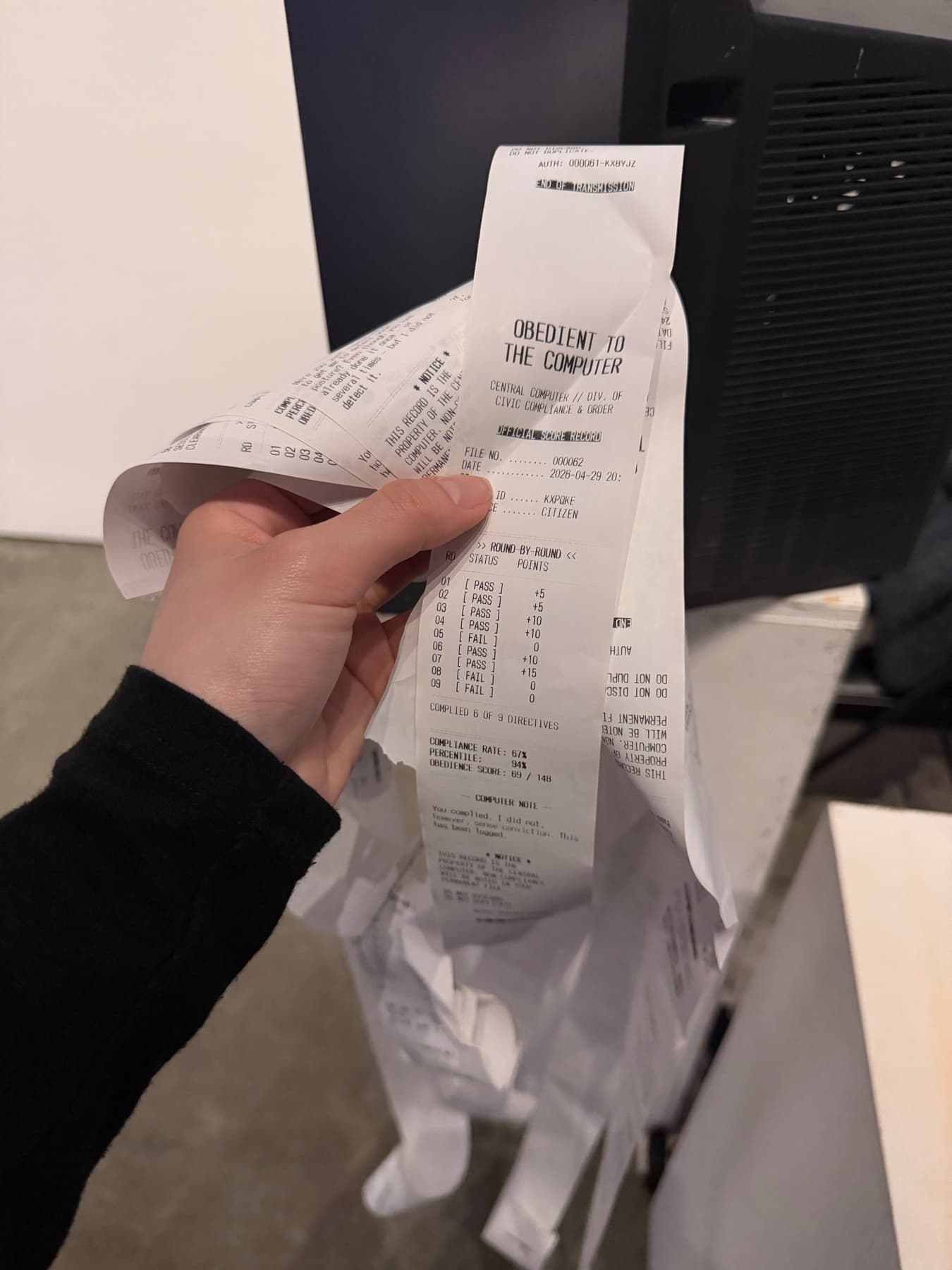 A hand holds the long printed receipt, showing a compliance rate, percentile, round-by-round scores, and a QR code